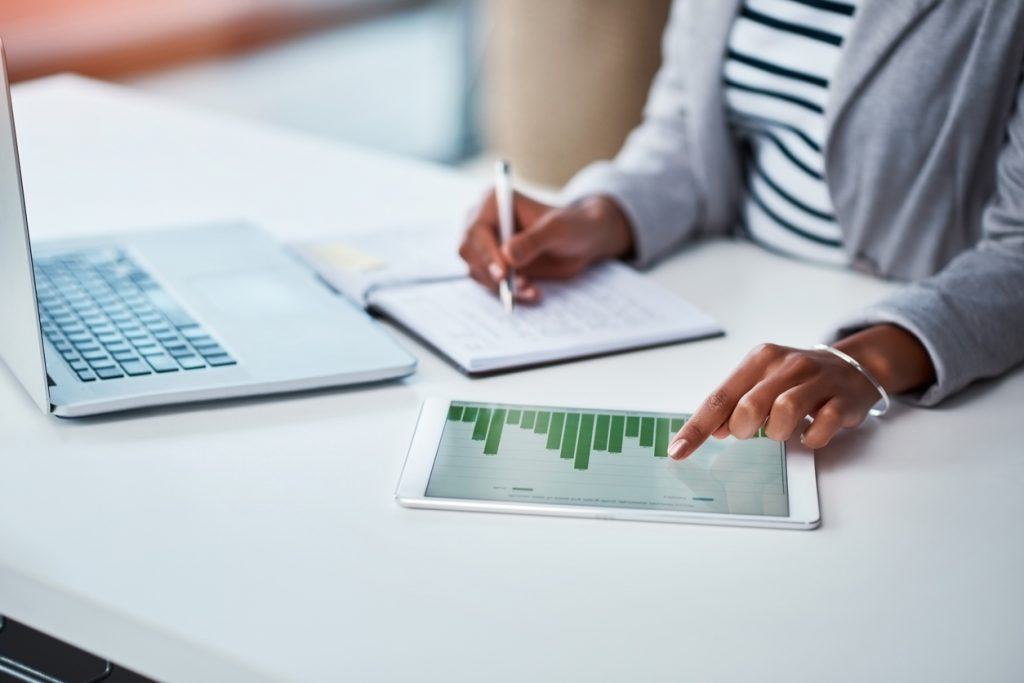 A customer experience manager sitting a desk with a laptop, tablet, and notebook doing data analysis.